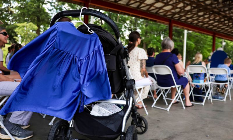 A blue child-sized graduation gown hangs from a stroller in the foreground of parents sitting at a Duke Children's Campus ceremony